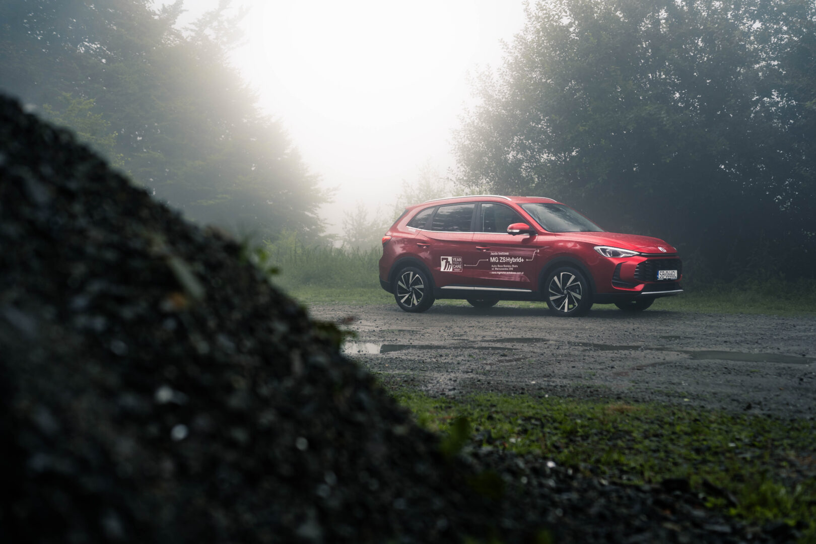 czerwony samochód MG ZS Hybrid+ stoi na parkingu w lesie, we mgle